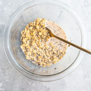 Mixed rolled oats with milk, chia seeds, vanilla and maple syrup in a clear mixing bowl with a wooden spoon.