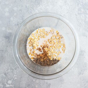Ingredients side by side in a clear mixing bowl: rolled oats with milk, chia seeds, vanilla and maple syrup.