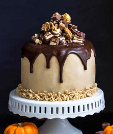 Side view of an entire buttercream frosted snickers cake with melted chocolate ganache on a white cake stand.