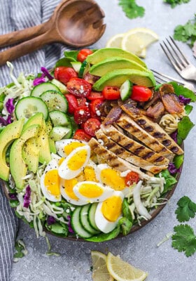 A bowl of Cobb salad on a countertop with a fork, fresh parsley and a wooden spoon beside it
