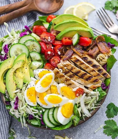 A bowl of Cobb salad on a countertop with a fork, fresh parsley and a wooden spoon beside it