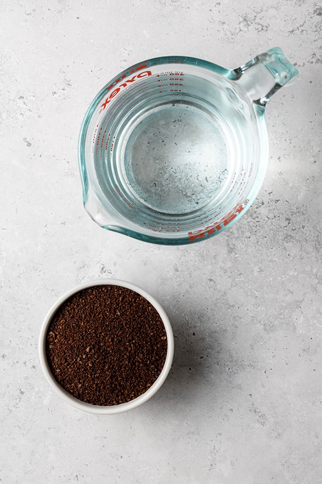 A measuring cup full of filtered water beside a bowl of coarsely ground coffee on a granite countertop