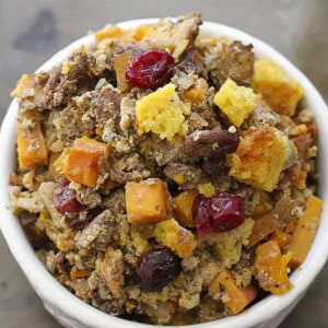A serving of cornbread stuffing with cranberries, butternut squash, apples and more in a white bowl on a brown background.