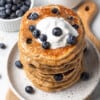 45 degree angle of a stack of gluten free blueberry pancakes topped with blueberries with whipped cream on a white plate