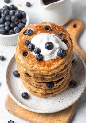45 degree angle of a stack of gluten free blueberry pancakes topped with blueberries with whipped cream on a white plate