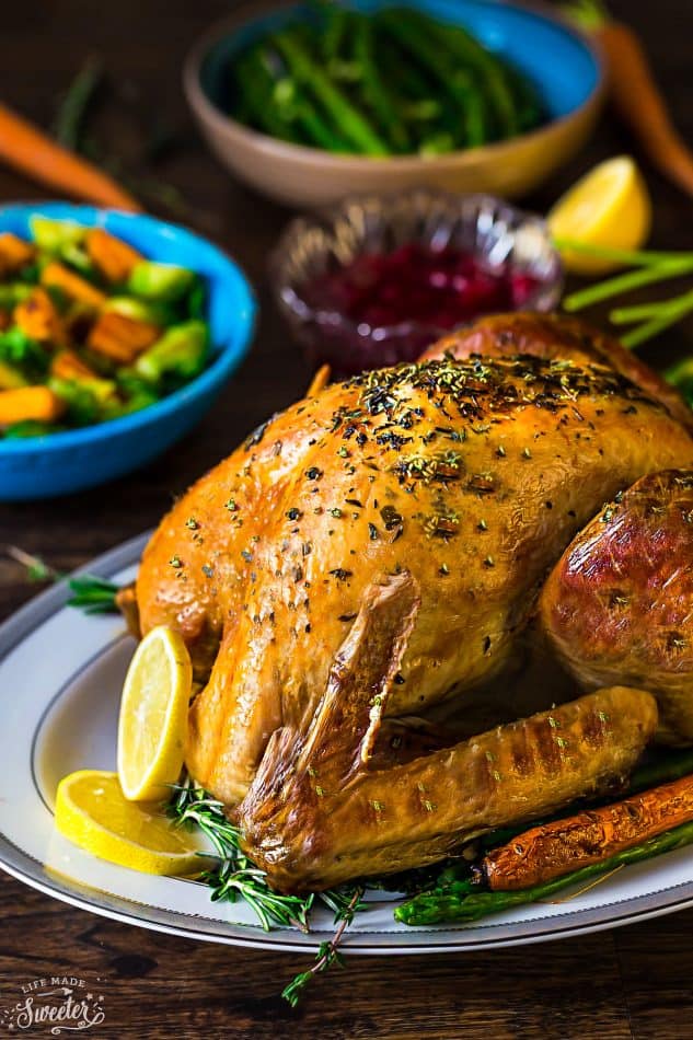 A roast turkey on top of a serving tray, garnished with herbs, and surrounded by lemon slices and fresh herbs