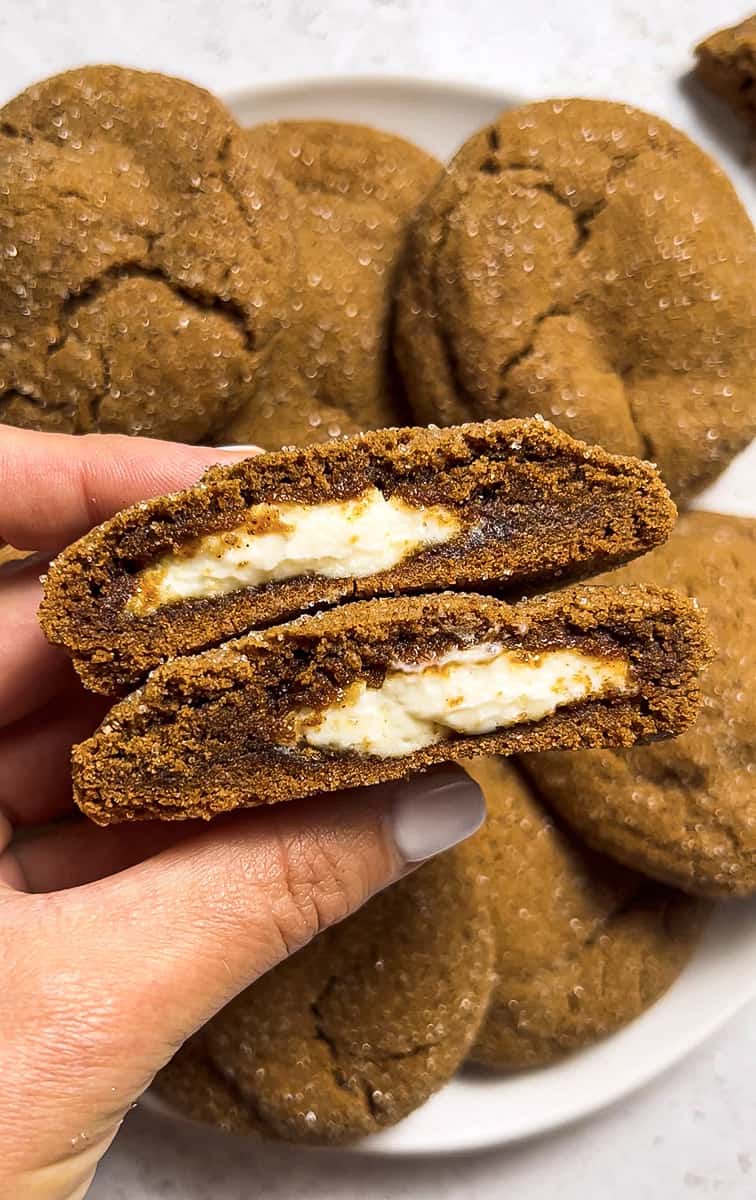 A hand holding two half gingerbread cream cookies over a white plate.