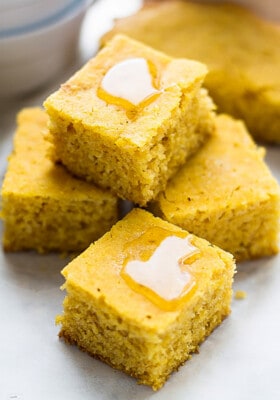 Four gluten-free cornbread squares topped with a drizzle of honey stacked on top in a pyramid on a white background.