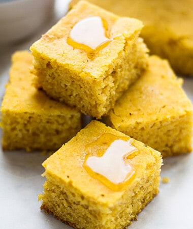 Four gluten-free cornbread squares topped with a drizzle of honey stacked on top in a pyramid on a white background.