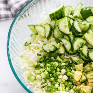 Chopped cabbage, green onions, cucumber and diced avocado in a large clear mixing bowl
