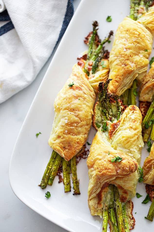 Close-up shot of a bunch of 4 of baked pastry asparagus bundles on a white rectangular platter