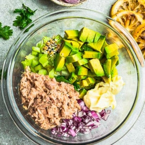 Top view of ingredients to make tuna salad in a clear bowl on a grey background with lemon, onions, celery, avocado, tuna and Whole30 mayo