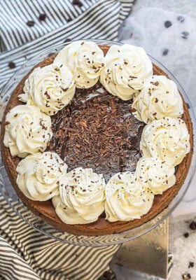 Top view of an entire Nutella Cheesecake on a clear cakestand with whipped cream and chocolate shavings on a grey background