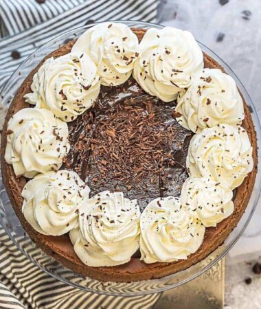 Top view of an entire Nutella Cheesecake on a clear cakestand with whipped cream and chocolate shavings on a grey background