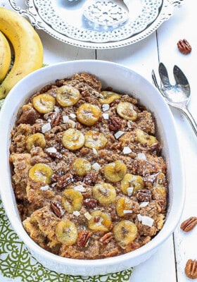 An entire banana french toast casserole in a white oval baking dish on a white background.