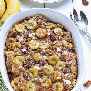An entire banana french toast casserole in a white oval baking dish on a white background.