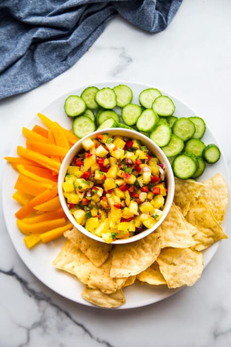 Top view of a bowl of fresh pineapple salsa served with sliced cucumbers, bell peppers and chips