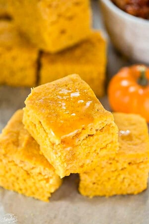 Three pumpkin cornbread squares topped with a drizzle of honey stacked on top in a pyramid on a white background.