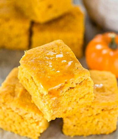 Three pumpkin cornbread squares topped with a drizzle of honey stacked on top in a pyramid on a white background.