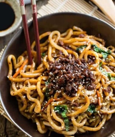 A bowl of Shanghai stir fried noodles topped with ground pork in a black bowl with chopsticks