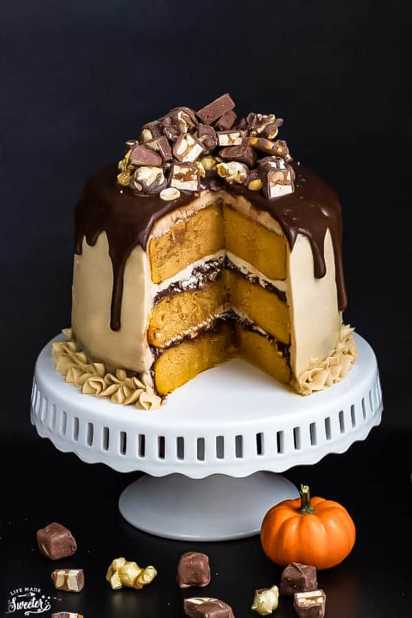 Side view of an entire buttercream frosted snickers cake with melted chocolate ganache cut in half on a white cake stand.