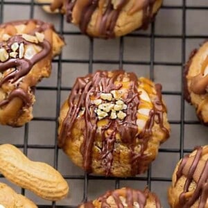 A close-up of a peanut snickers cookie surrounded by 5 other cookies drizzled with melted chocolate and caramel sauce on a black wire rack.