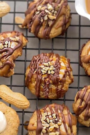A close-up of a peanut snickers cookie surrounded by 5 other cookies drizzled with melted chocolate and caramel sauce on a black wire rack.