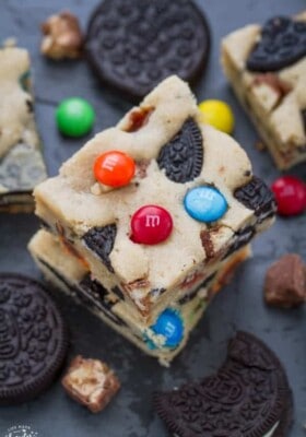 Two Monster Cookie Bars stacked on top of each other on a black baking sheet surrounded by Oreo cookies and M&M's