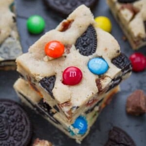 Two Monster Cookie Bars stacked on top of each other on a black baking sheet surrounded by Oreo cookies and M&M's
