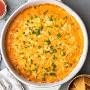 A batch of vegan buffalo chicken dip in a white round baking dish with a serving of chips on the side