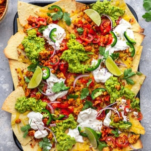 Overhead shot of loaded vegan nachos layered with seasoned tofu crumble, guacamole, vegan sour cream and fresh tomato salsa in a baking pan on a grey background