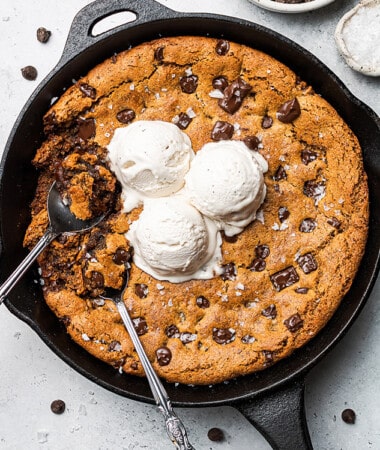 A giant chocolate chip cookie inside of a cast-iron skillet with scoops of dairy-free vanilla ice cream on top