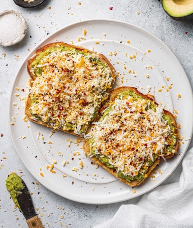 Top shot of two slices of toasted bread with mashed avocado and grated hard boiled egg, salt, black pepper and red pepper chili flakes on top on a white plate