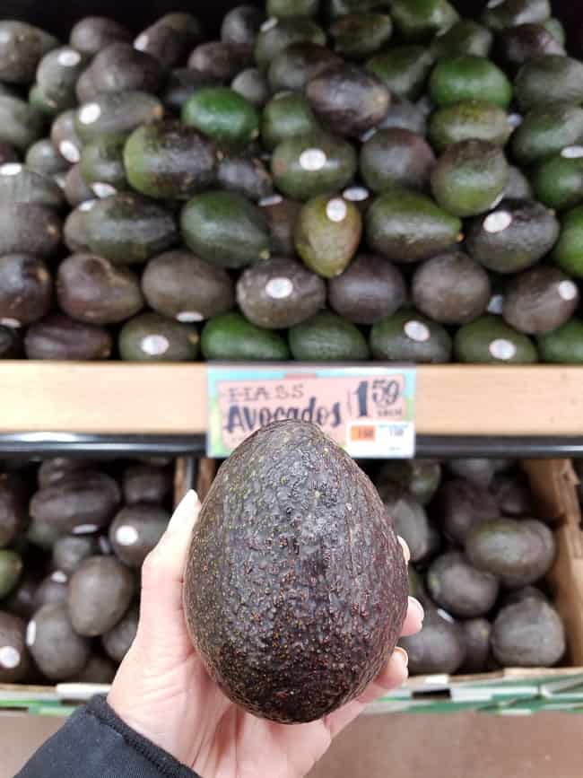 A hand holding an Avocado in front of avocados in a bin at the grocery store