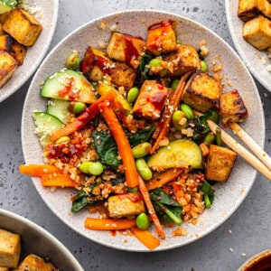 A mixed bowl of vegan bibimbap in a white bowl with crispy tofu, matchstick carrots, edamame, cucumber, spinach and red cabbage together with gochujang sauce and riced cauliflower with chopsticks
