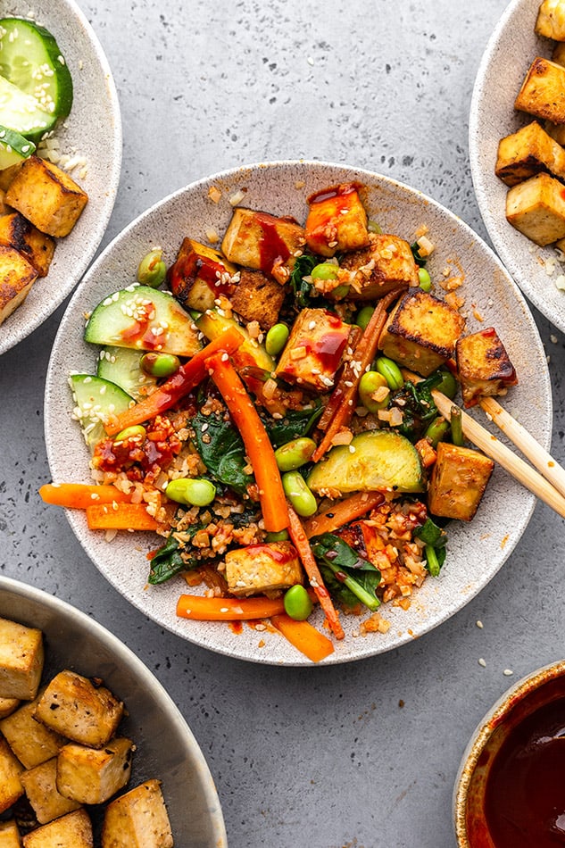 A mixed bowl of vegan bibimbap in a white bowl with crispy tofu, matchstick carrots, edamame, cucumber, spinach and red cabbage together with gochujang sauce and riced cauliflower with chopsticks