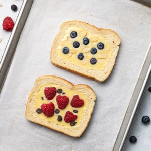 Two slices of untoasted custard toast topped with raspberries, blueberries, almonds and chocolate chips on a baking sheet