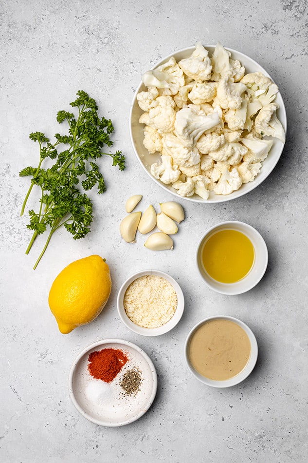 A bowl of cut cauliflower, six garlic cloves, a lemon and the rest of the ingredients on a granite countertop