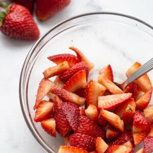 A clear mixing bowl of sliced strawberries with a spoon