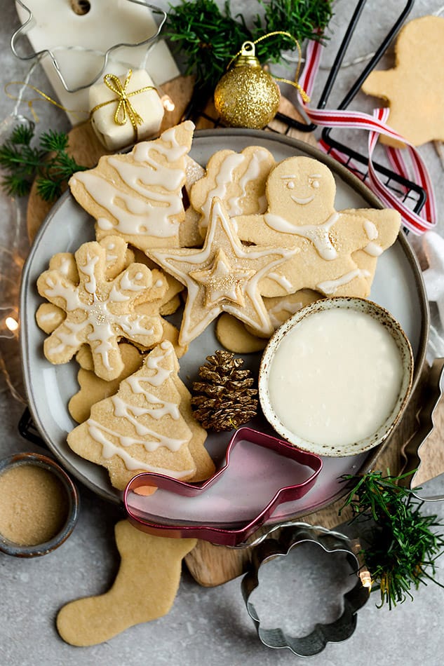 A plate with decorated sugar cookies and a bowl of icing, surrounded by cookie cutters and Christmas decorations