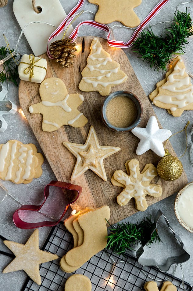 A cutting board with decorated sugar cookies, surrounded by cookie cutters and Christmas decorations