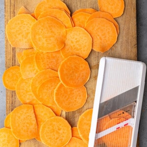 Sliced raw sweet potato rounds on a wooden cutting board beside a mandolin