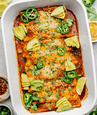 A pan of veggie enchiladas in a white casserole pan topped with avocado, cilantro, jalapeno and dairy-free cheese.