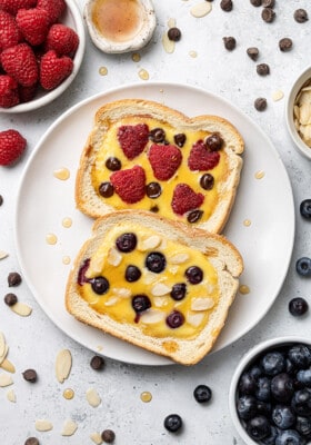 Top shot of two slices of toasted custard toast topped with raspberries, blueberries, almonds and chocolate chips on a white plate
