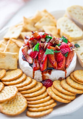 Side shot of a pile of Balsamic Strawberries piled on whpped feta served with an assortment of gluten-free crackers and toasted baguettes