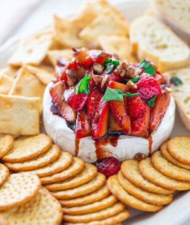 Side shot of a pile of Balsamic Strawberries piled on whpped feta served with an assortment of gluten-free crackers and toasted baguettes