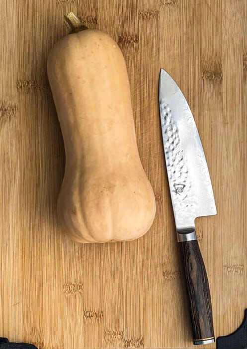A whole unpeeled butternut squash on a wooden cutting board beside a sharp knife