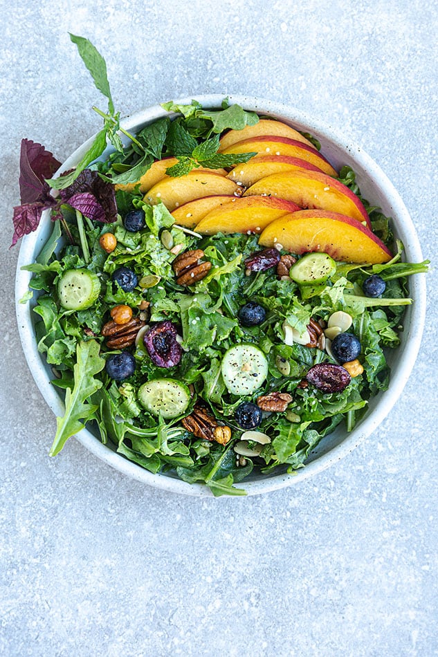 Top view of arugula salad with peaches and blueberries in a white bowl on a grey background