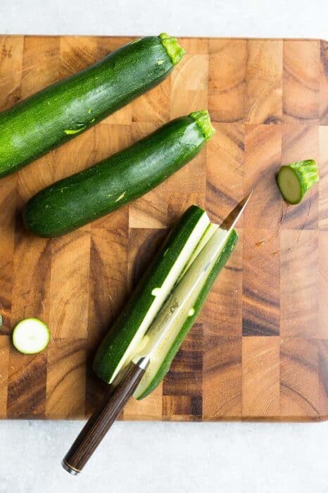 An uncooked zucchini being sliced in half on a wooden cutting board
