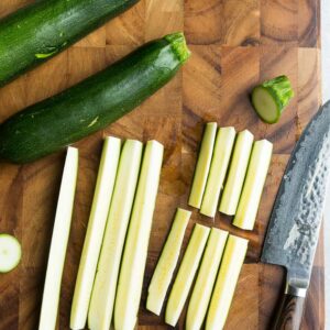 Raw zucchini spears on a cutting board with a sharp knife and two full zucchini